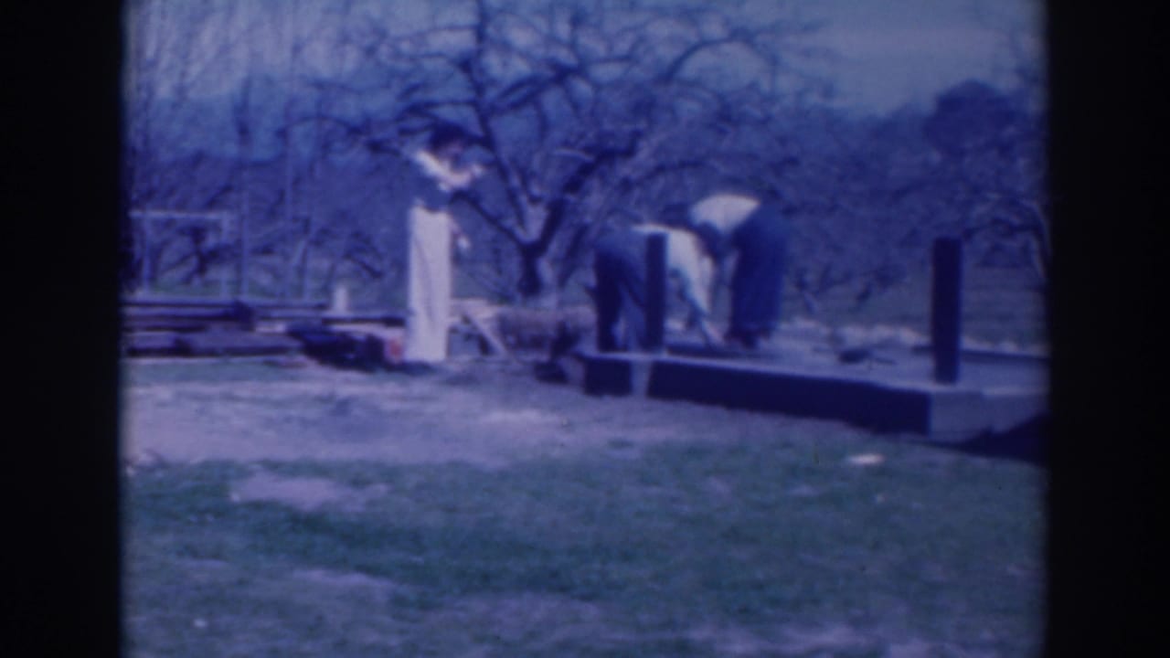Adults work near a small platform with upright posts in a grassy yard.