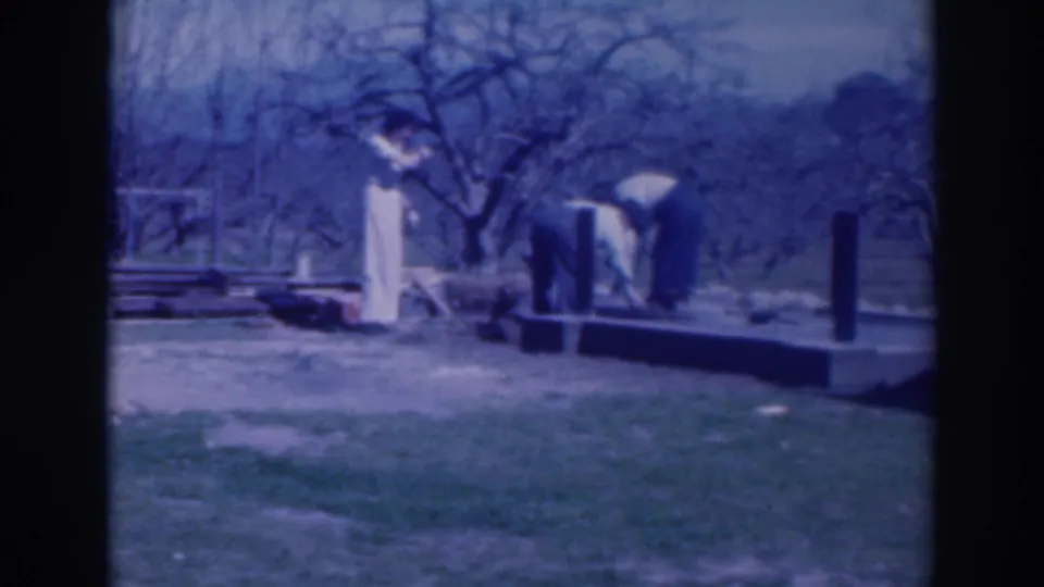 1937 Sacramento home-movie frame showing adults around a low platform with posts, beams, grass, and bare trees.