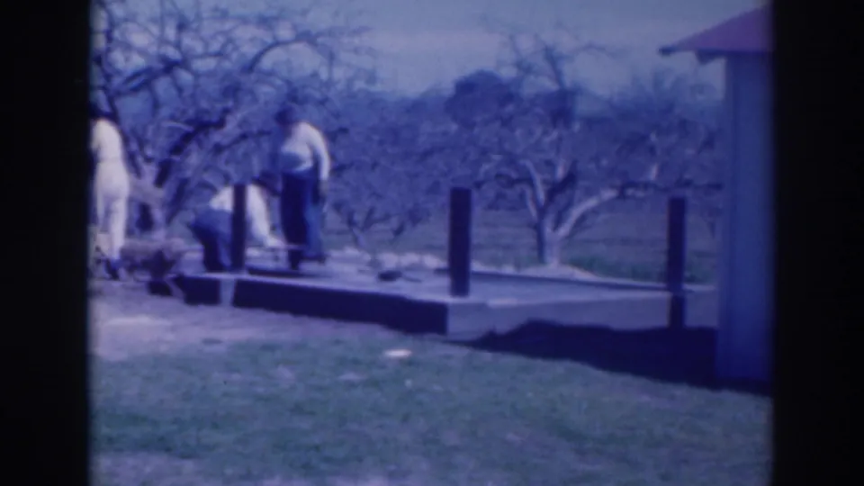 1937 Sacramento home-movie frame showing adults near a yard platform, lawn, bare trees, and a light structure at the edge of frame.