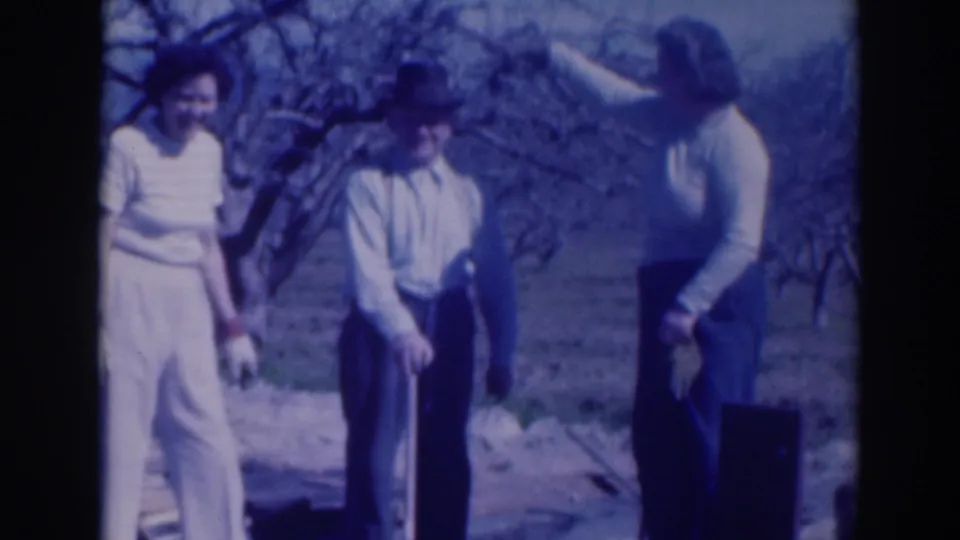 In this 1937 Sacramento home-movie frame, three adults stand in a garden or orchard area, the man wearing a fedora and suspenders.