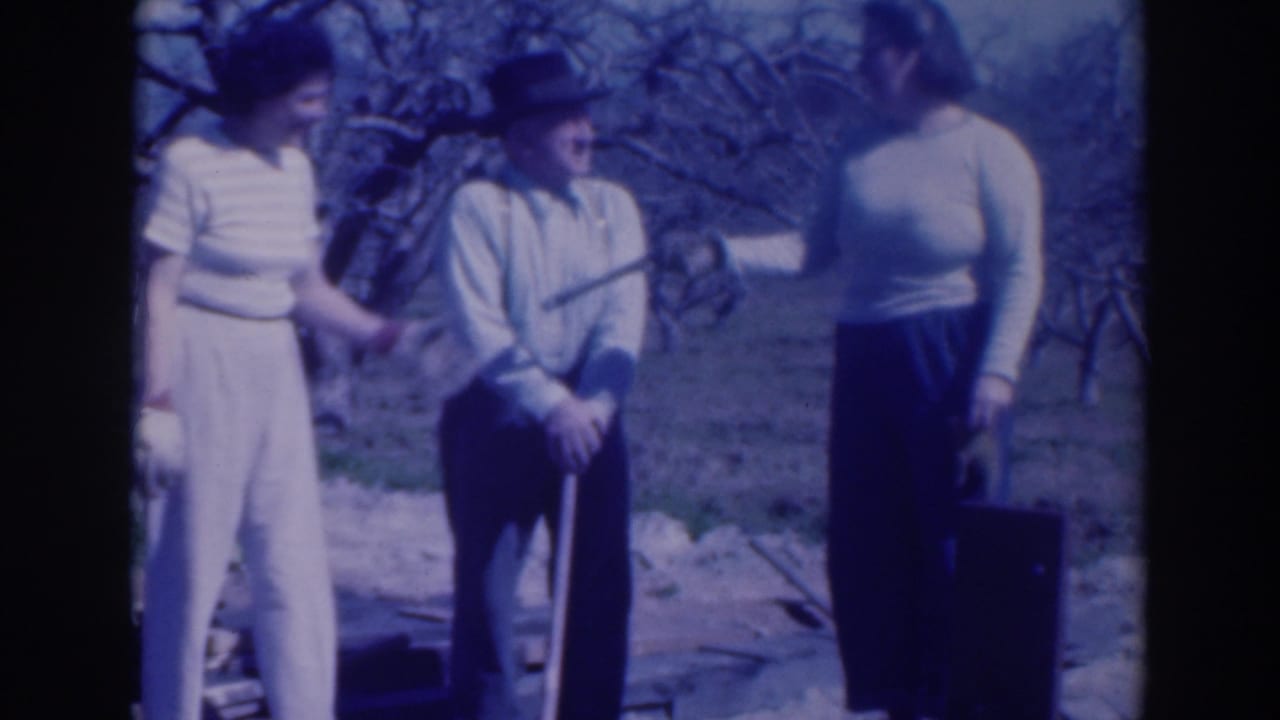 In a 1937 Sacramento orchard scene, two women stand close to an older man in a hat as he holds a long-handled tool.