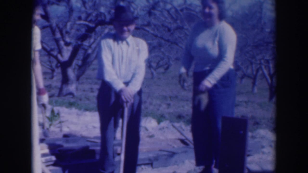 Adults stand outdoors near a shovel and bare trees during a small dig scene.