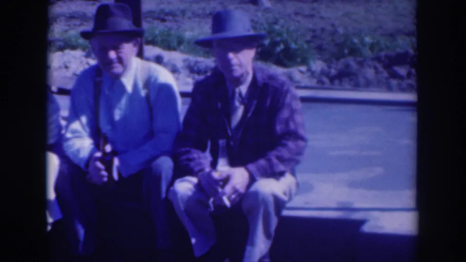 Two hat-wearing men sit side by side in the 1937 Sacramento home movie, one holding a bottle near a paved outdoor area.