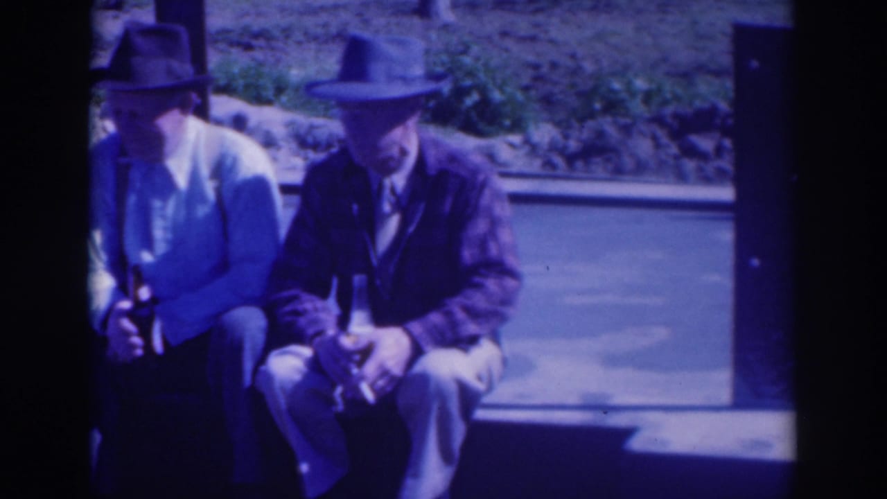 Two men in hats sit on a bench holding bottles beside a road.
