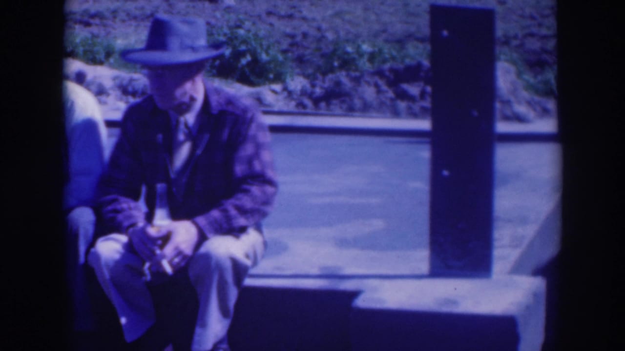 A seated man in a fedora holds a bottle while another man is partly visible.