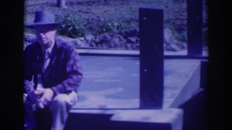 A single seated man in a fedora and jacket holds a bottle near a road and vertical post in blue-tinted 8mm home-movie footage.