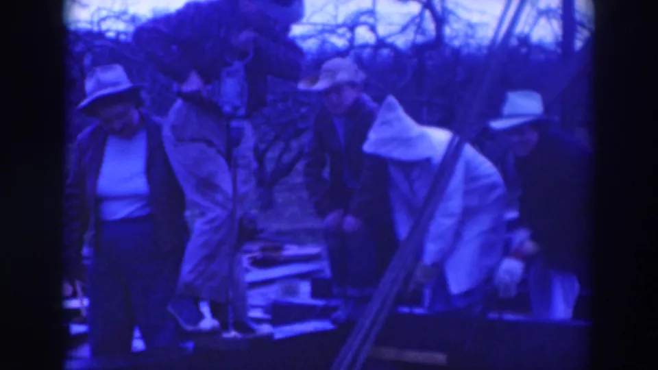 1937 home-movie frame from Sacramento showing adults clustered around a pit or equipment in a bare wooded outdoor area.