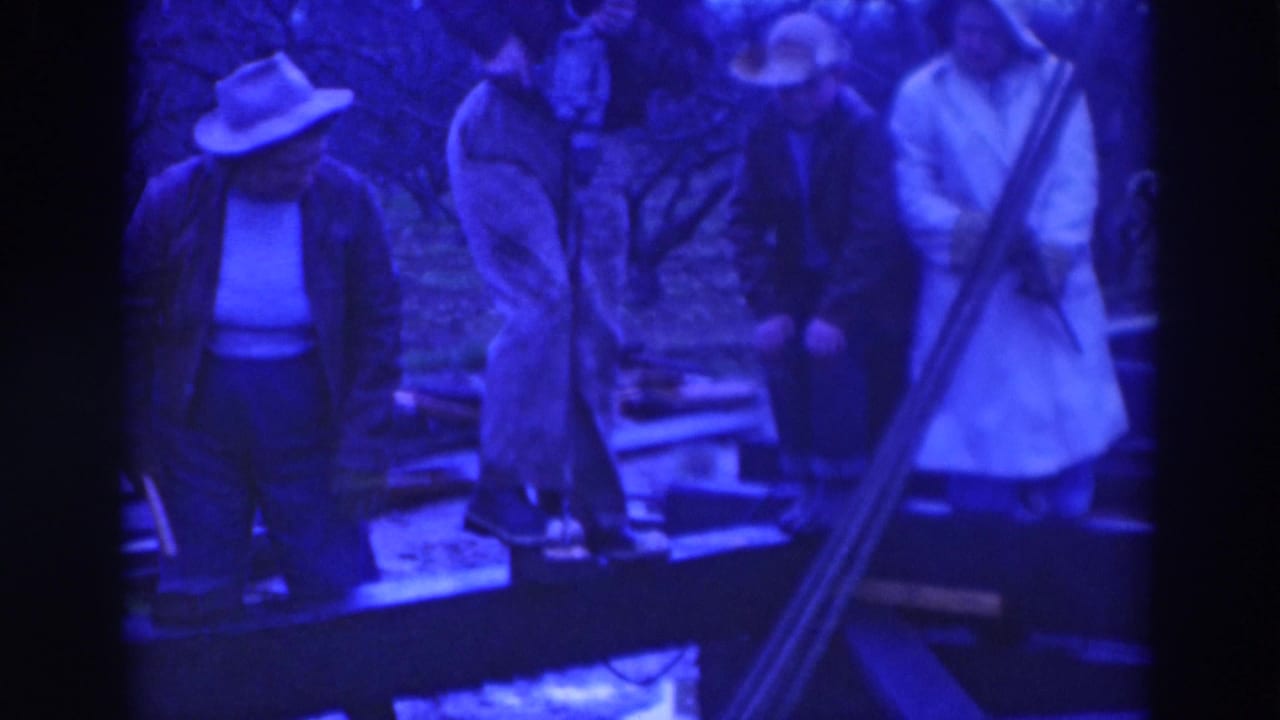 Adults in hats stand near a wooden platform and boards in a blue-tinted outdoor construction scene.