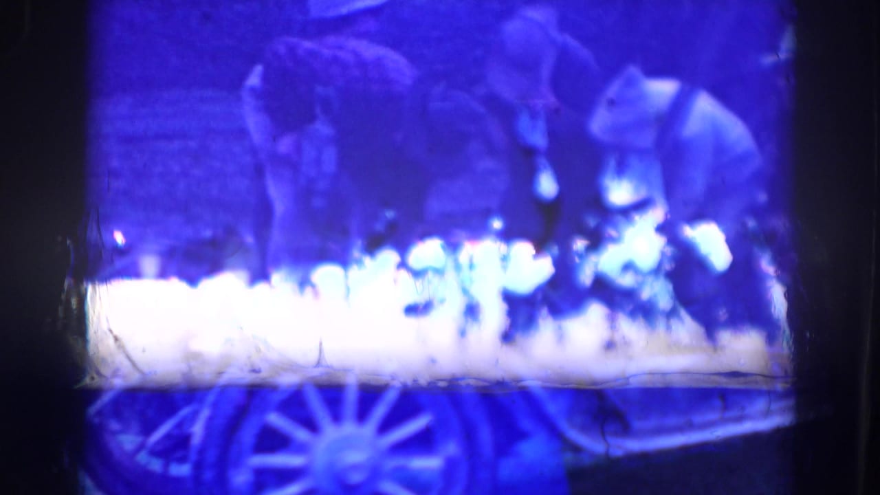 Blue-tinted film frame with blurred figures near a spoked-wheel automobile.
