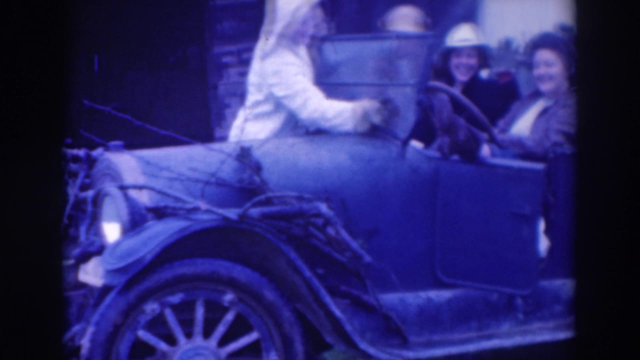 Women seated in an open-top car while another person stands beside it.