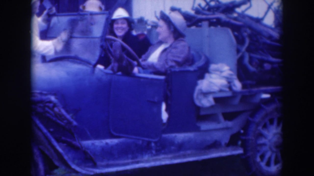 Two women in hats sit in an open vintage car with firewood stacked behind them.