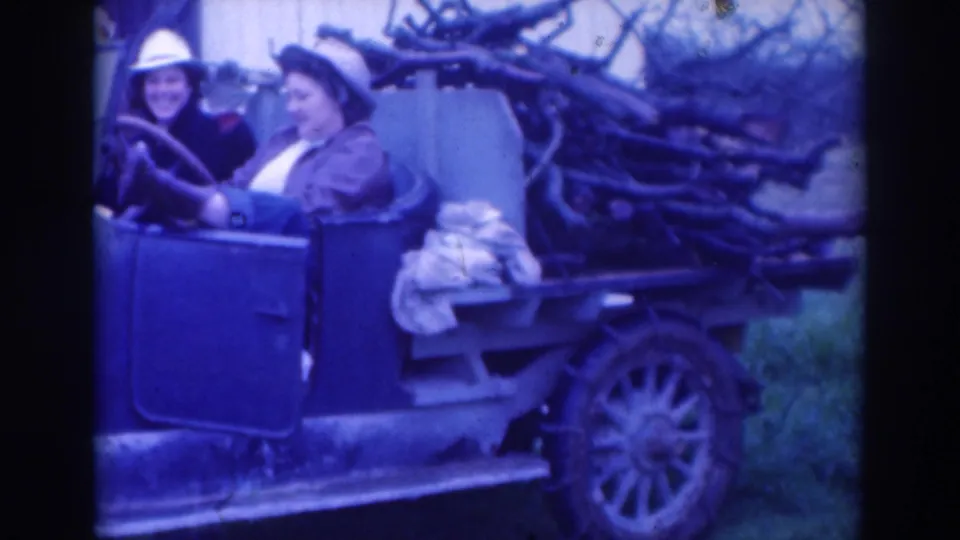 A driver and passenger in hats smile in a 1937 Sacramento home movie, seated in an open vintage car with firewood behind them.