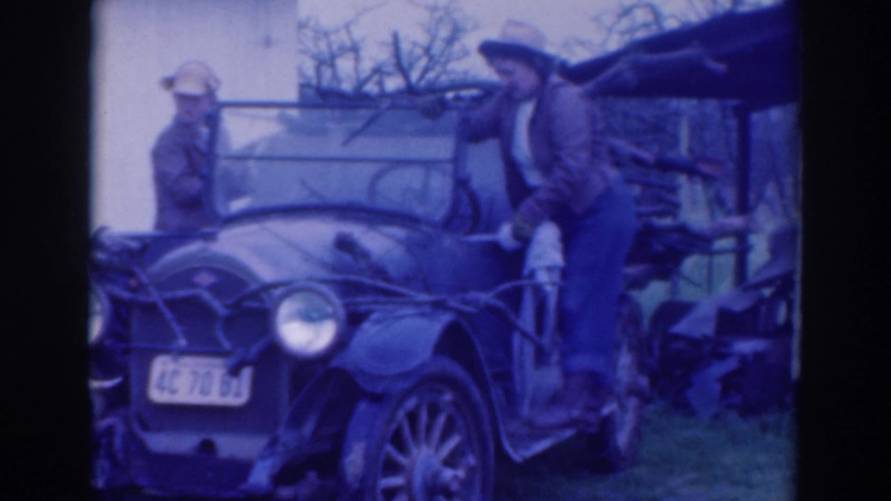 A man stands on the running board of a vintage car near the windshield as another man watches.