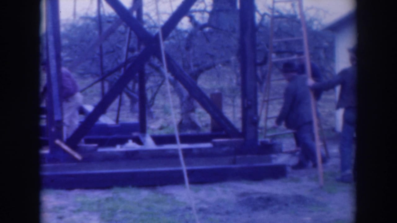 Workers stand near a large wooden framework with ladders and lumber outdoors.