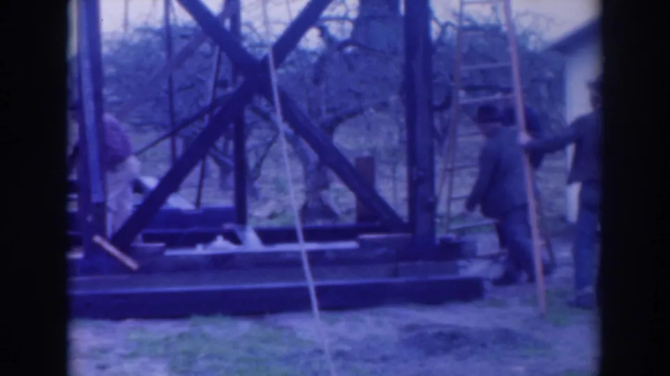 1937 Sacramento home-movie frame showing workers around a large wooden structure with ladders, braces, and bare trees behind it.