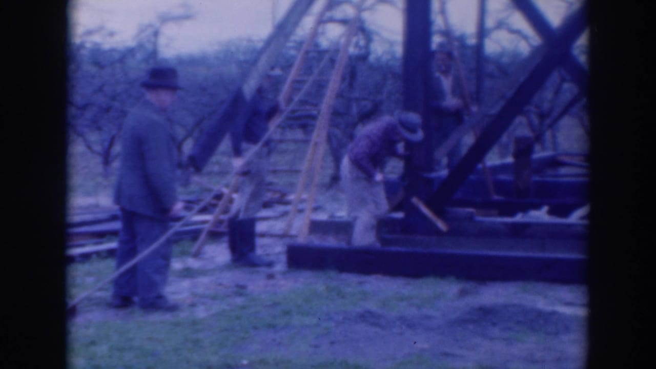 Men work beside a raised wooden frame with ladders, ropes, grass, and bare trees visible in the background.