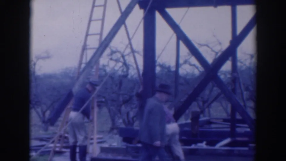 Workers are seen beneath tall wooden supports, with a ladder, rope, grass, and leafless trees around the site.