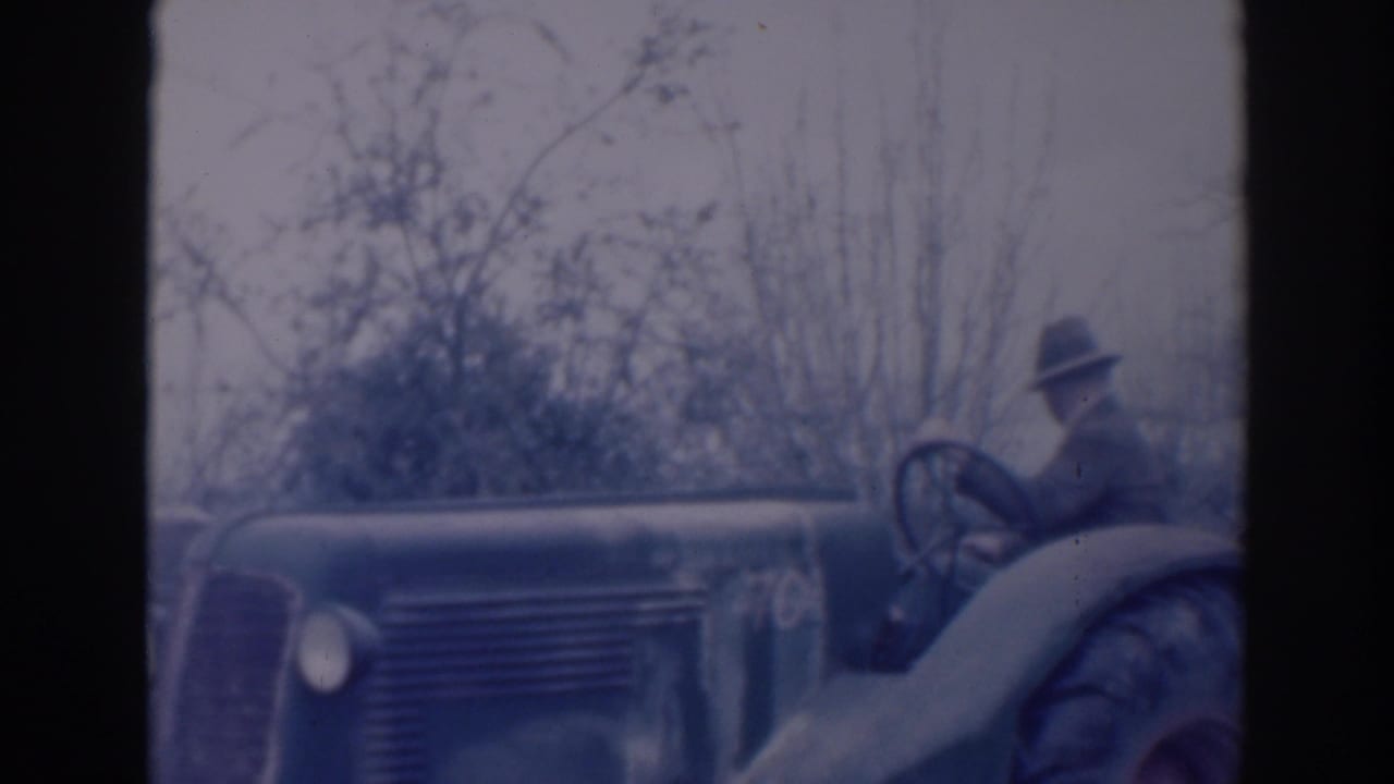Older man in a brimmed hat seated on a tractor beside bare trees.