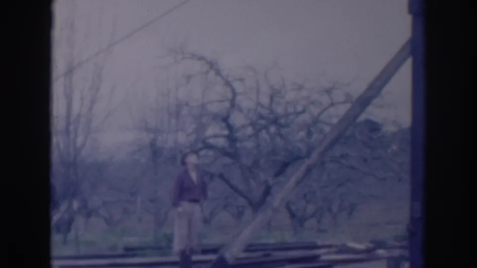 In 1937 Sacramento home-movie footage, a man stands near diagonal construction framing with leafless trees and an overcast sky behind him.
