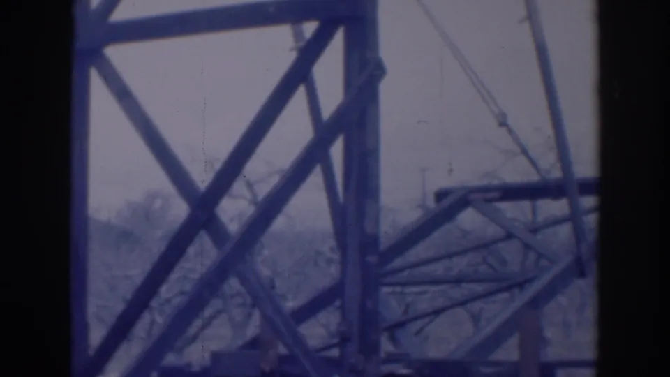 1937 Sacramento home-movie frame with a steel grid, diagonal supports, suspended cables, and bare trees seen through the structure.