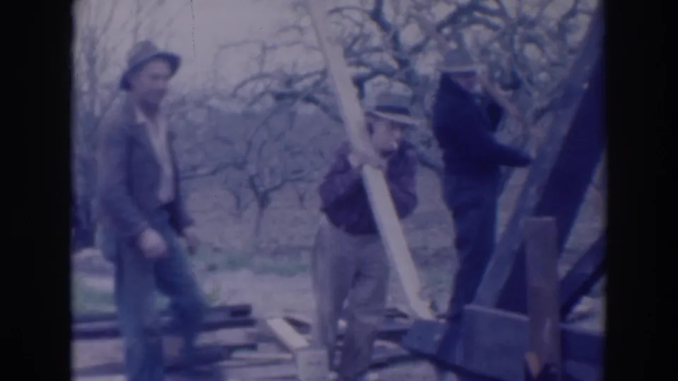 Two men in hats stand beside a large angled wooden beam in the Sacramento orchard scene, with lumber visible on the ground.