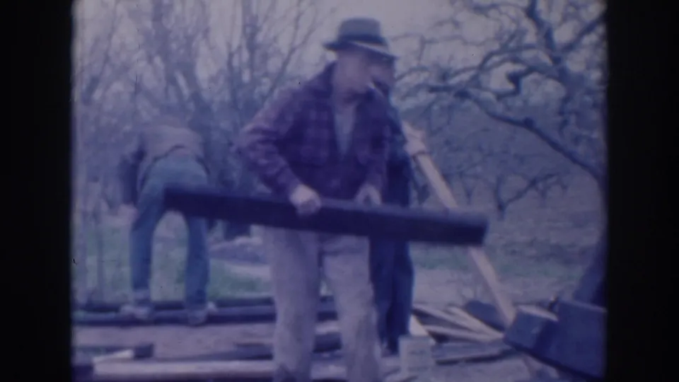 A man in plaid work clothing lifts a long board as two other men continue outdoor work in the 1937 Sacramento scene.