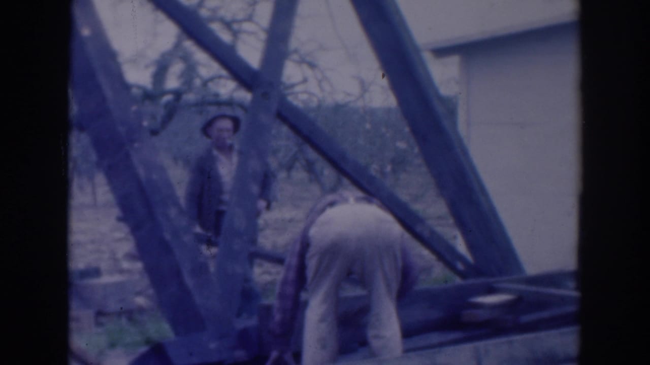 Two adult men near a wooden structure outdoors, one standing in a jacket and hat while another bends forward.