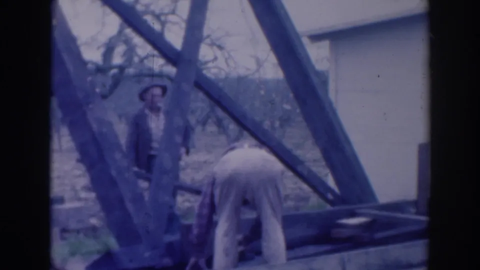 Outdoor 1937 Sacramento home-movie frame with one man observing beside a wooden structure and another working bent over in light trousers.