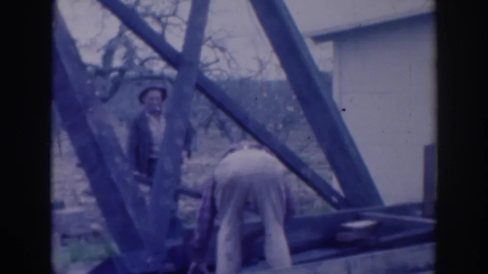 Blue-tinted 8mm frame from Sacramento in 1937 showing two men beside a wooden structure, bare trees, and a pale exterior wall.