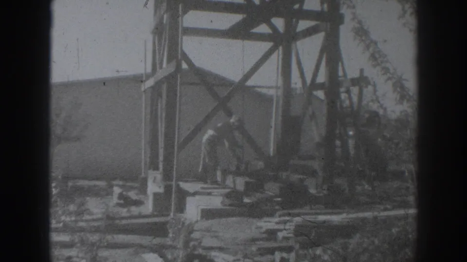 1937 Sacramento frame with a construction worker under a timber derrick structure near cables, braces, and building materials.