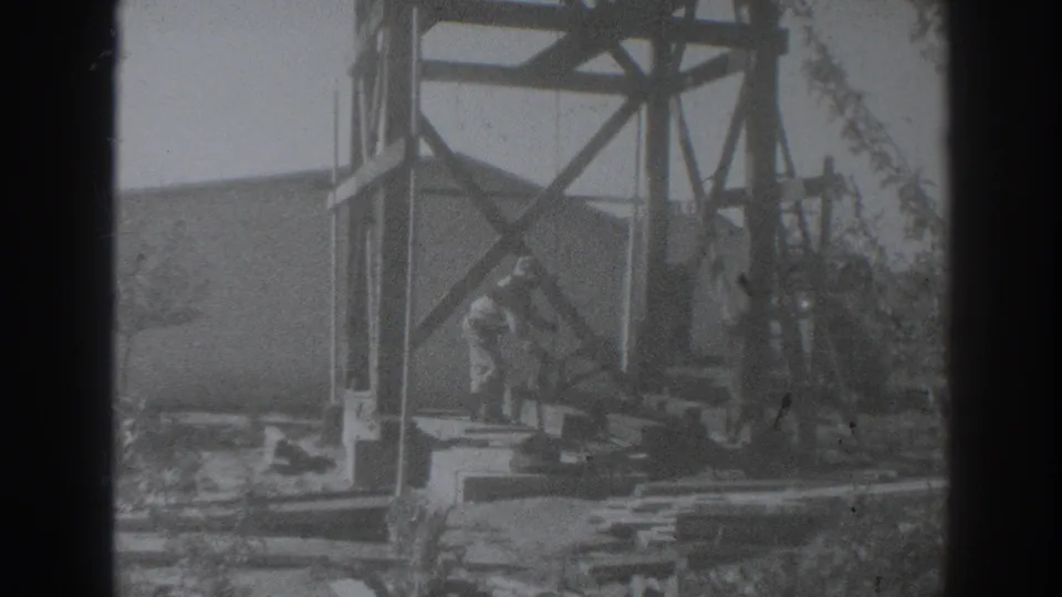 1937 Sacramento home-movie frame showing a worker handling equipment near the base of a wooden derrick at a building site.