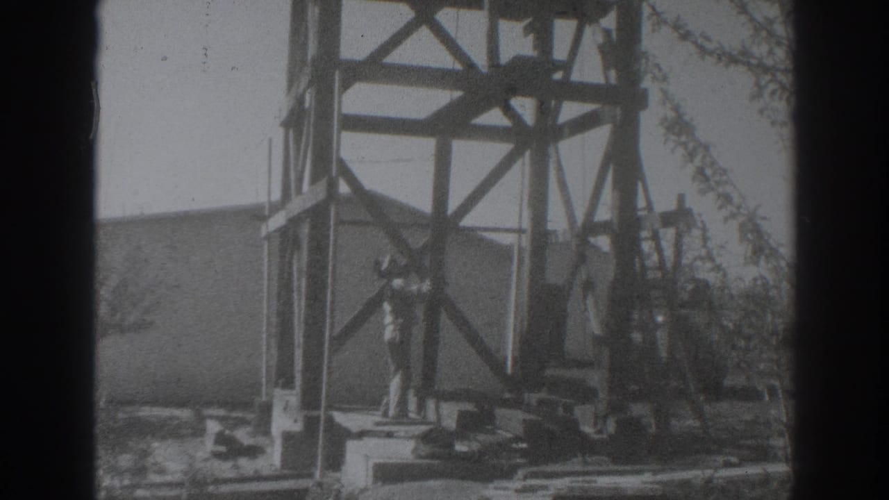 Man stands beside a wooden tower frame with lumber and a low building behind him.
