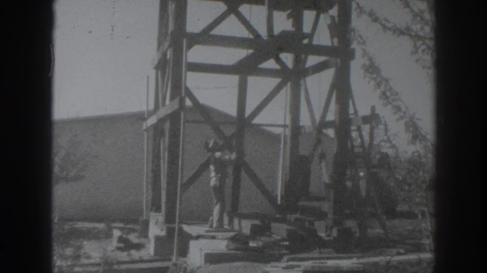 In 1937 Sacramento home-movie footage, a man works near a wooden tower frame with boards and a plain building wall behind him.