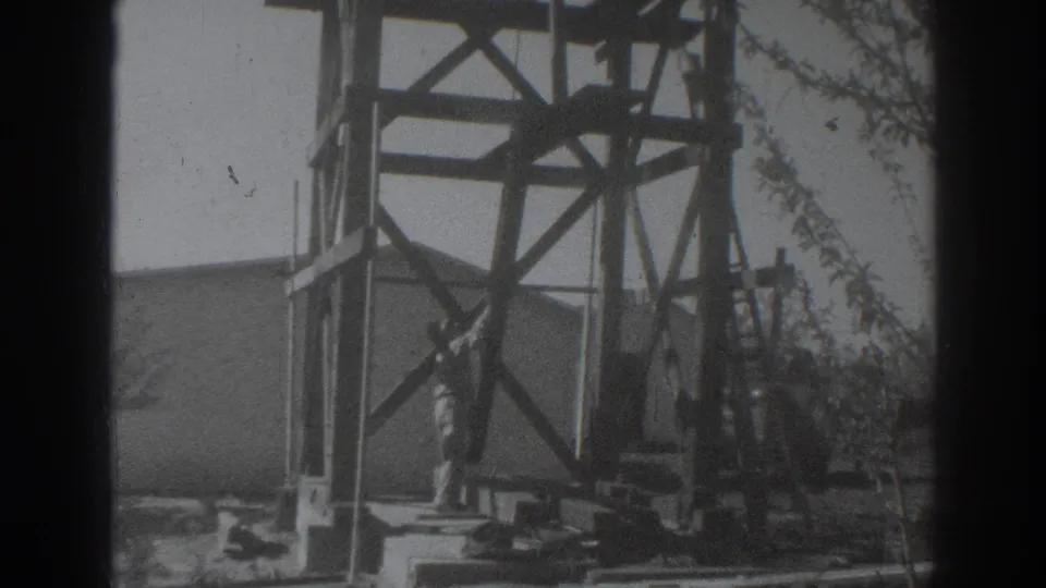 The 1937 home-movie frame shows the man working low near the base of the wooden tower, with lumber scattered in the foreground.