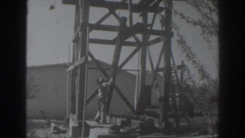 The man appears beside the wooden tower frame in Sacramento, surrounded by boards, a plain wall, branches, and visible 8mm film marks.