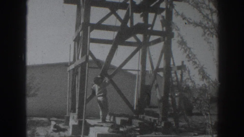 The man continues positioning a long beam beside the timber framework, with a plain building and trees behind the work area.