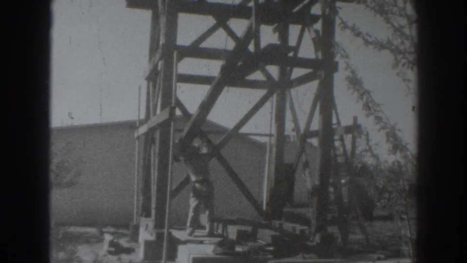 Near the end of the clip, the worker bends at the base of the wooden structure as the 8mm frame shows heavy grain and edge shadows.
