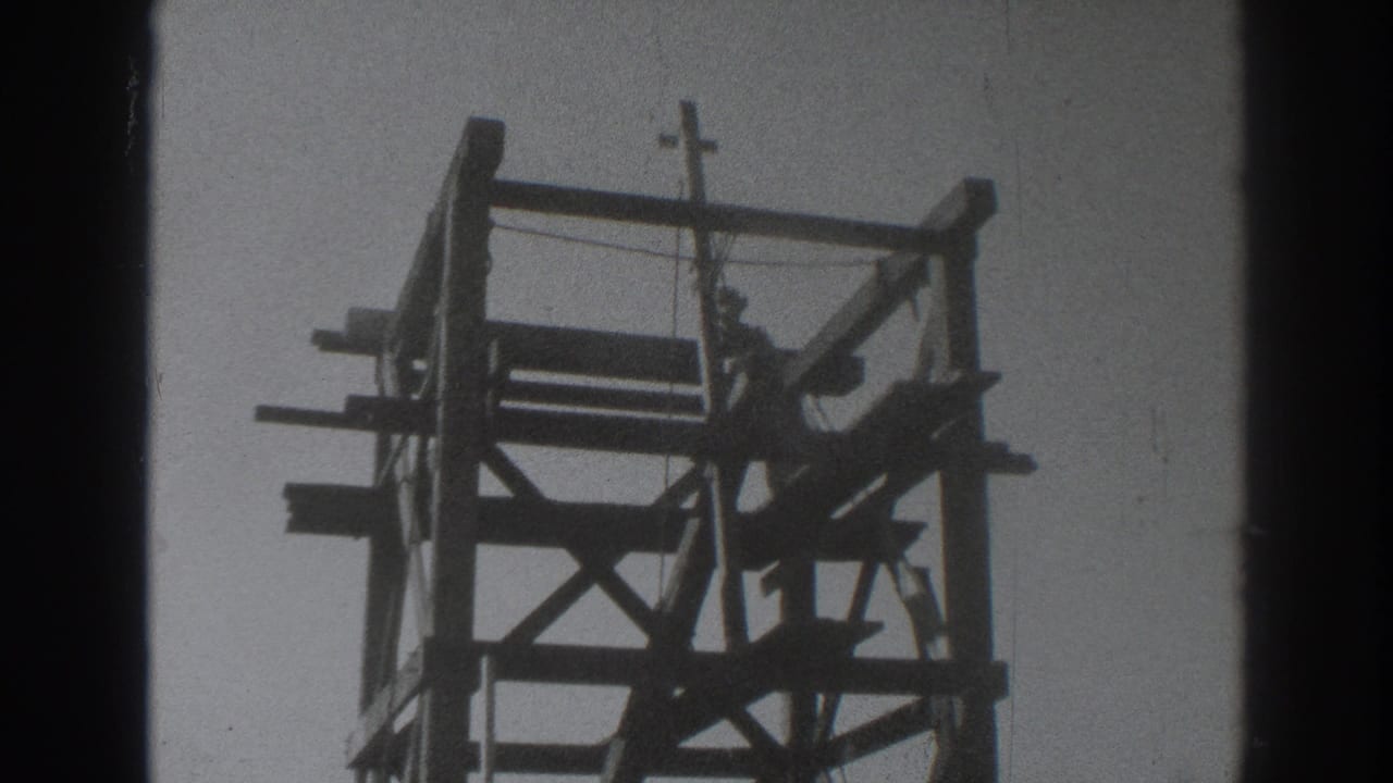 Silhouetted man working near the top of a wooden tower with a crucifix above.