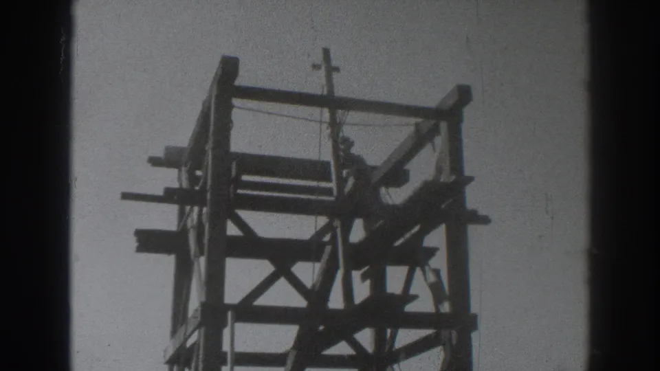In 1937 Sacramento footage, a man appears silhouetted on a wooden tower frame beneath a small crucifix against a bright sky.