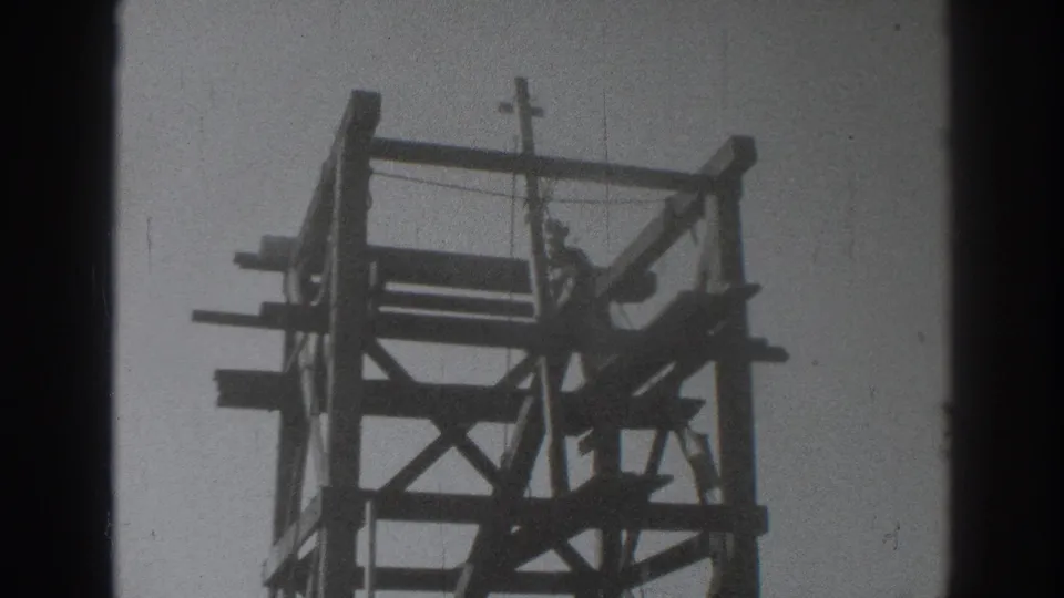 The 1937 Sacramento frame shows the worker within the upper bracing of a wooden tower, with the crucifix visible above him.