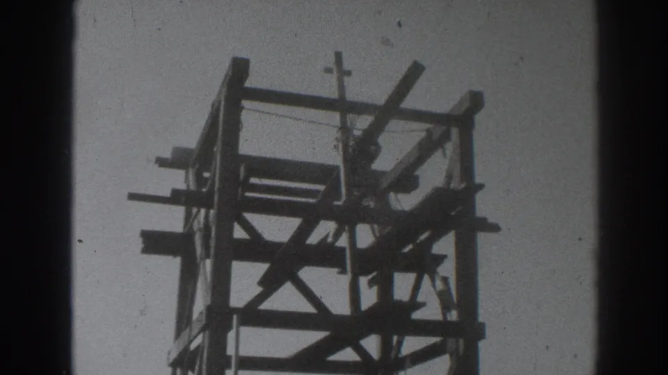 1937 Sacramento home-movie frame of a worker high in a timber tower structure, with a small cross visible near the top.