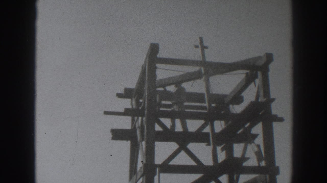Man working near the top of a wooden tower with a small crucifix above.