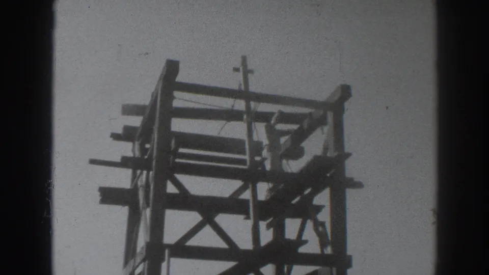 Low-angle 1937 home-movie view of the wood rig frame, showing X bracing, platform beams, and bright sky around the structure.