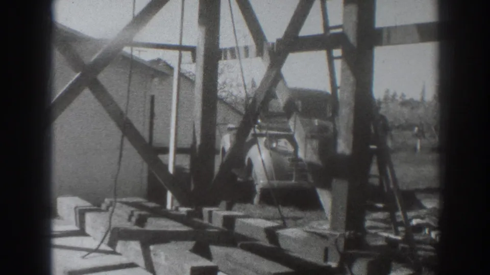 Black-and-white 8mm frame of a rural Sacramento construction area with timber supports, boards, and a period car in the background.