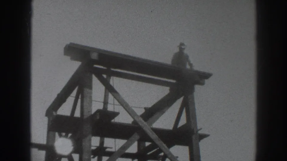 1937 Sacramento frame showing a worker on the upper edge of a wooden scaffold, viewed from a low angle against the sky.
