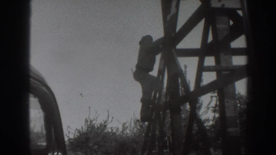 1937 Sacramento frame showing a worker on the side of wooden scaffolding, with brush near the base and pale sky behind.