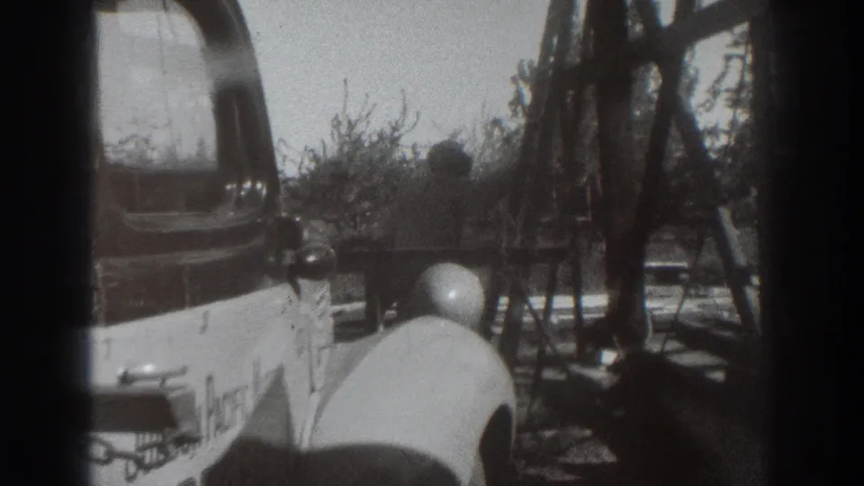 Near the end of the clip, the man appears closer to the base of the wooden structure beside the car and shrubs.