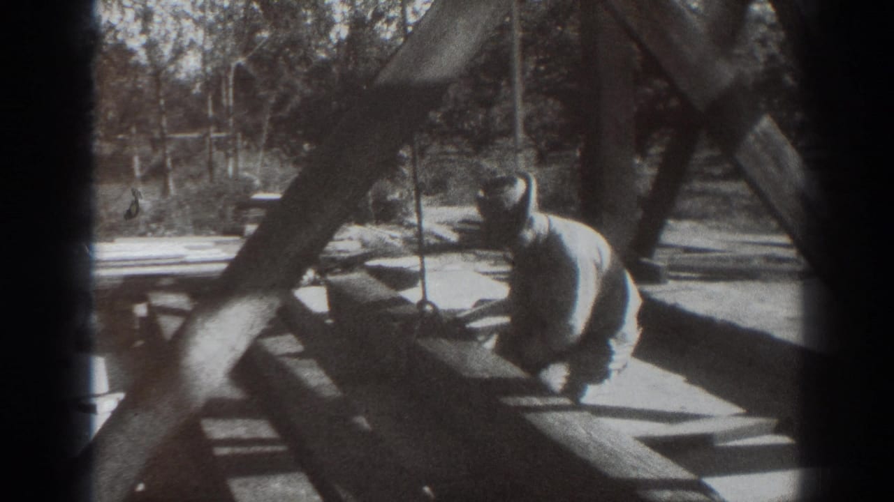 Man kneeling beside a wooden framework outdoors with trees behind him.