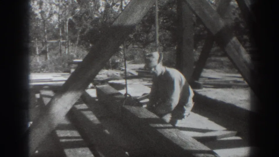A man sits or kneels near angled timber supports, with trees and fencing visible in the background.