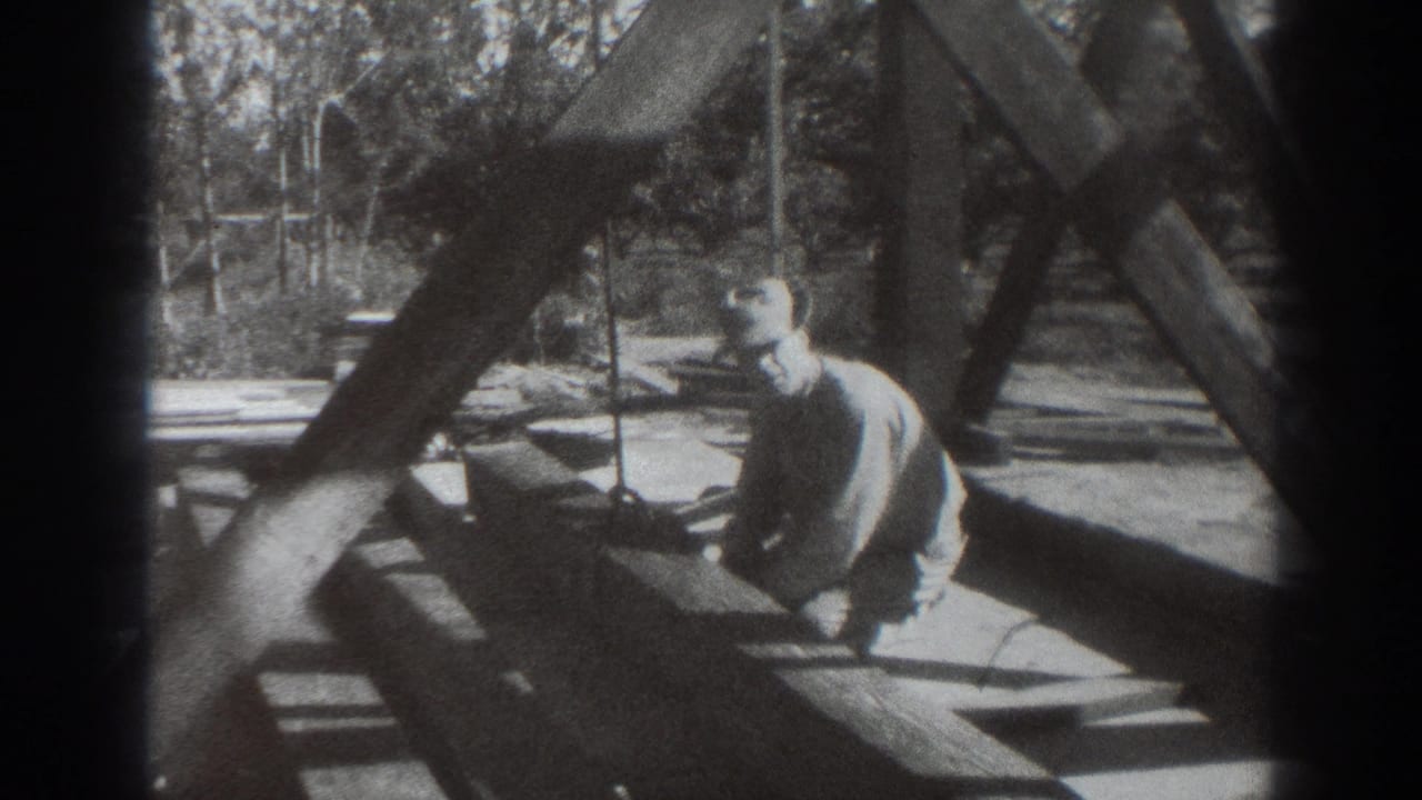 Worker kneels beside large wooden beams and handles rope in an outdoor structure.
